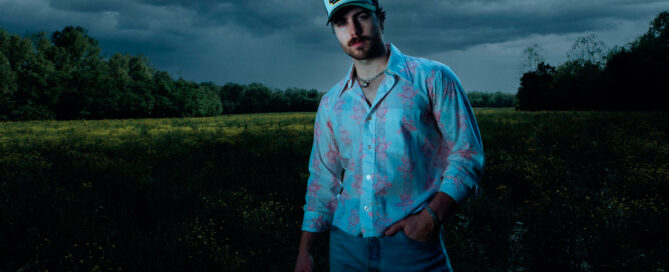 Young man standing in a field on a cloudy day, wearing a blue floral shirt, jeans, and a cap.