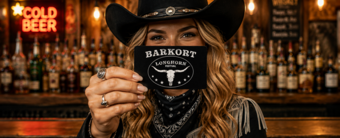 Woman in a black cowboy hat holds up a black card that reads 'BARKORT Longhorn Festival' in a bar setting with warm, blurred bottles in the background.