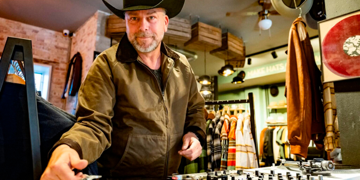 Man wearing a cowboy hat and brown jacket operates a DJ mixer in a clothing store venue.