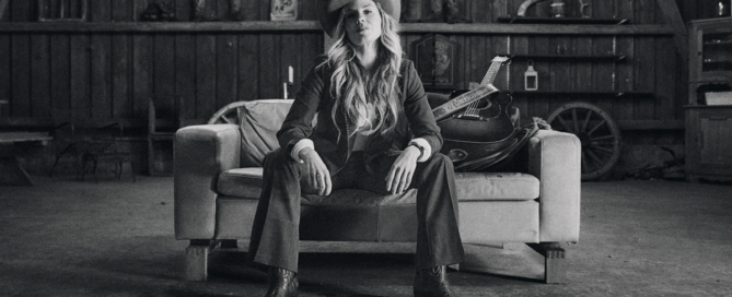 Woman wearing a cowboy hat sits on a sofa in a rustic workshop with a guitar resting beside her, looking at the camera (black-and-white).