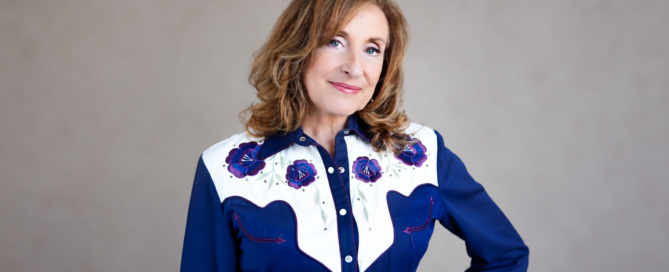 Portrait of a smiling woman with wavy light brown hair wearing a navy blue Western-style shirt with a white yoke and purple floral embroidery.